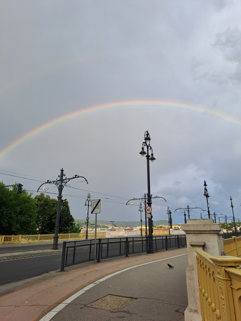 Photo on a rainbow over Margaret Bridge in Budapest, early on a cloudy Sunday morning. The bridge is empty except for a wandering pigeon. The full arc of the rainbow arches over the elaborate lampposts and balustrades.