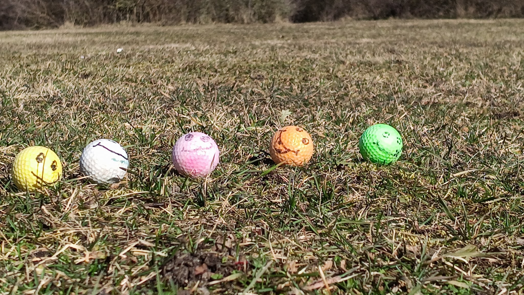 Auf einer noch grauen Wiese liegen bunte Golfbälle, die alle ein auf gemaltes Gesicht haben und uns anlächeln