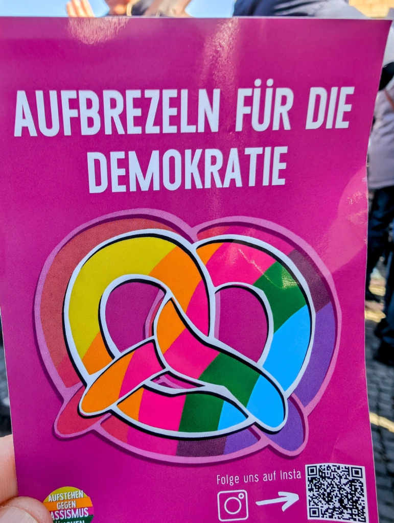 Das Bild zeigt eine pinkfarbene Broschüre oder ein Schild, das von einer Person gehalten wird. Oben auf dem Schild steht in großen, weißen Buchstaben der Slogan "AUFBREZELN FÜR DIE DEMOKRATIE". Darunter ist eine stilisierte Brezel abgebildet, die in verschiedenen Regenbogenfarben wie Gelb, Orange, Pink, Grün und Blau dargestellt ist. Am unteren Rand des Schildes befindet sich der Text "Folge uns auf Insta" zusammen mit einem Instagram-Logo und einem QR-Code. In der unteren linken Ecke ist ein kleines Logo mit dem Text "AUFSTEHEN GEGEN RASSISMUS MÜNCHEN" zu sehen. Der Hintergrund des Bildes ist unscharf, aber es sind Menschen und Umrisse von Gebäuden zu erkennen, was darauf hindeutet, dass das Foto im Freien aufgenommen wurde.