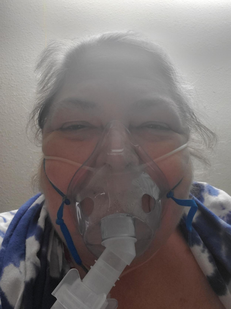 Me with a nebulizer mask on, vaporizing medicine for my lungs, dark blond hair in a ponytail, blue and white shawl draped over the shoulders