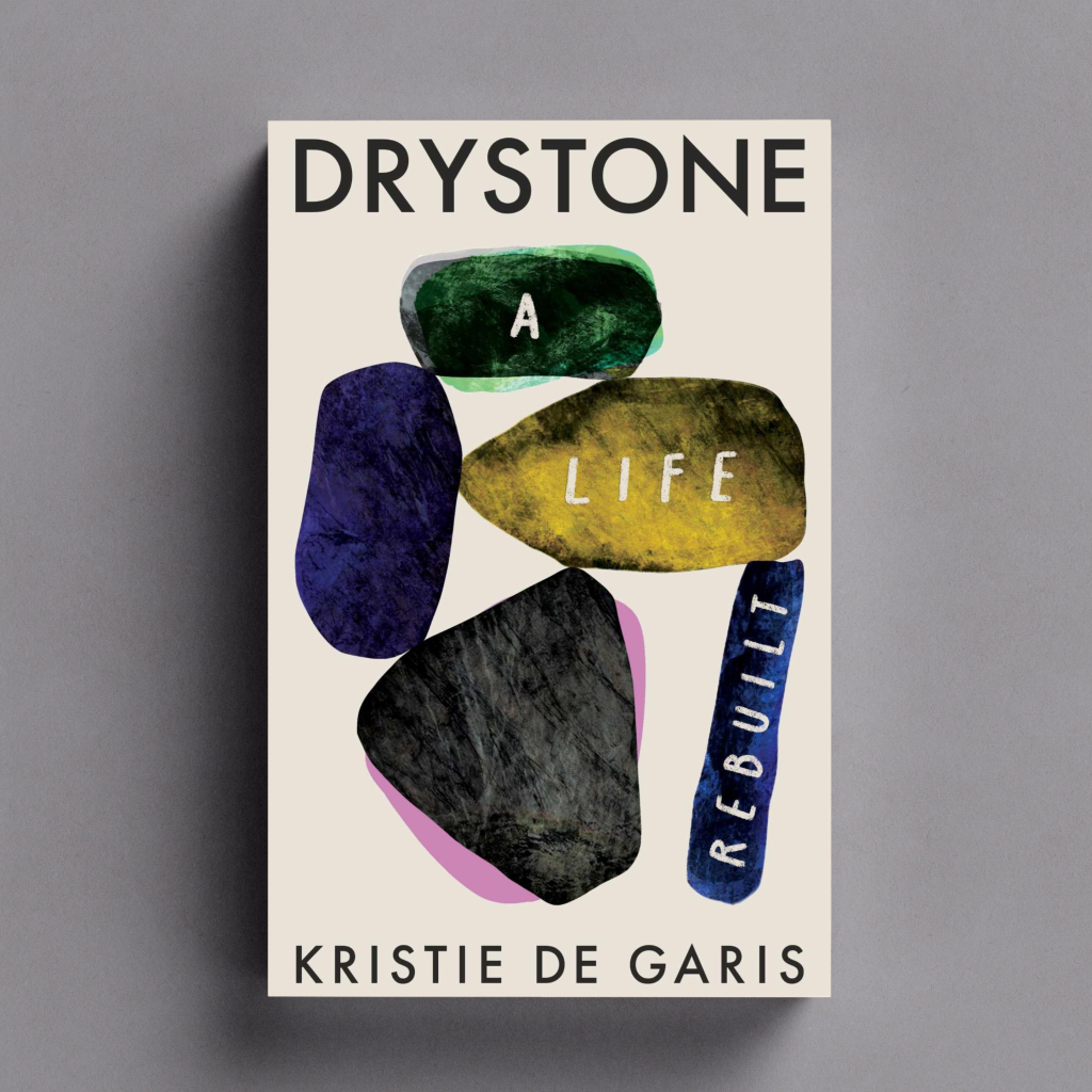 A book titled 'Drystone - A Life Rebuilt'. It's cover has 5 large colourful stones on it. At the top a green one which leans on a purple and a ochre stone which lean on a blue and a slate grey stone with a pink background. On the green, yellow and blue stones the words 'A Life Rebuilt' are written. 