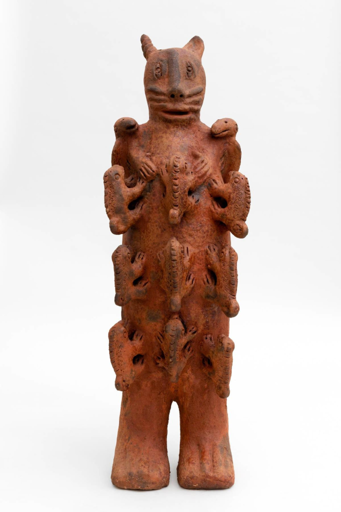 Freestanding red clay sculpture of a figure with one catlike head on top and three tiers of iguana-like creatures perched along their body in neat rows.