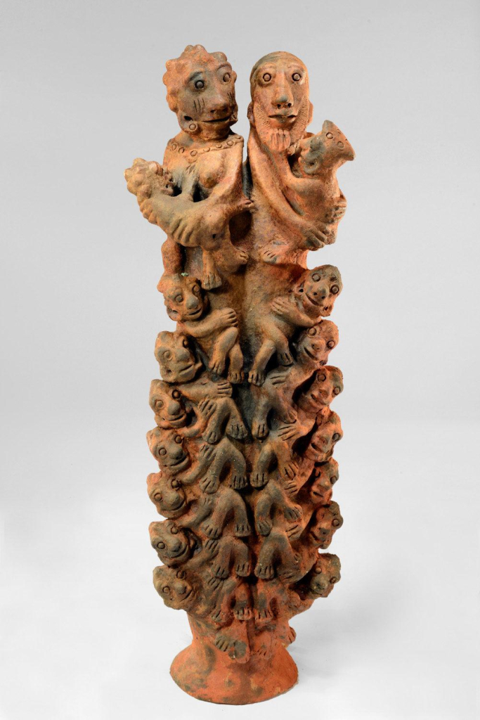 Freestanding red clay sculpture two figures on top each holding a smaller infant creature, and many vertical layers of smaller smiling creatures stacked along the body atop one shared base at the bottom.