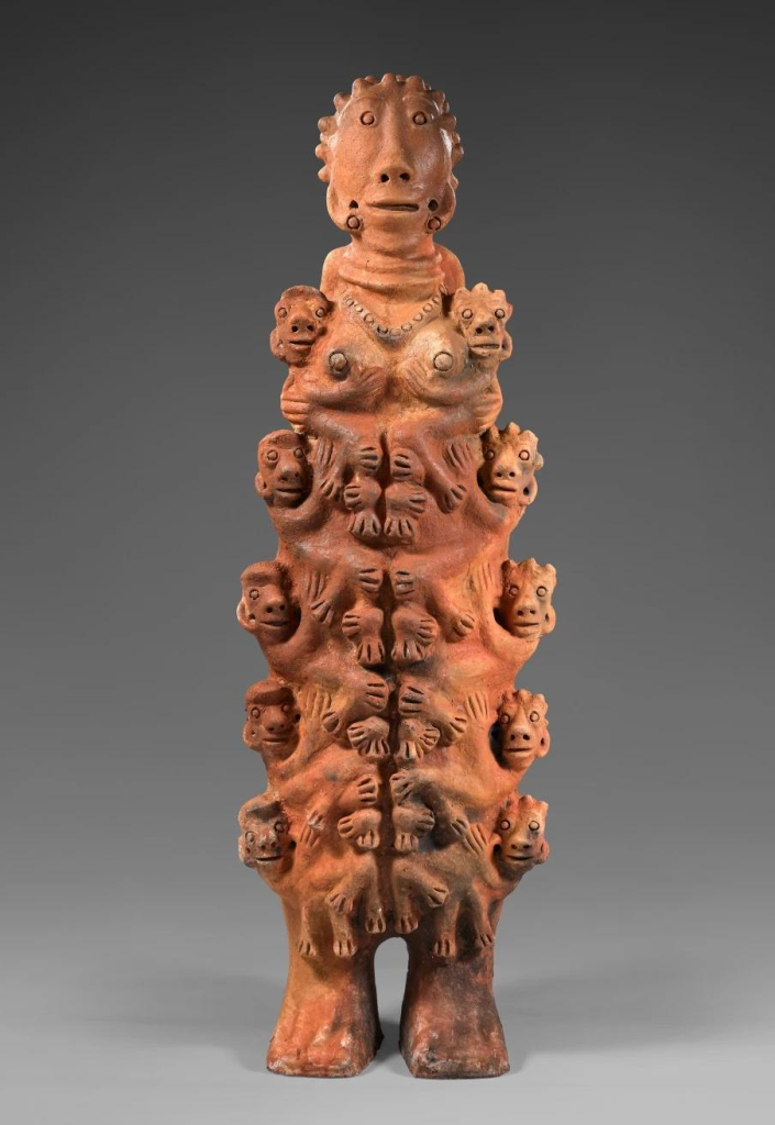 Freestanding red clay sculpture of a figure with one smiling head on top and several tiers of smiling creatures hugging their body along both the right and left sides.