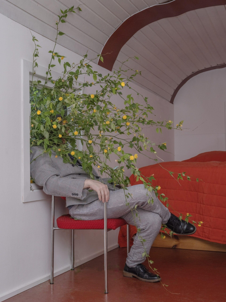 Photo of a white figure in a grey suit sitting on a red chair in a small white room with a rounded ceiling and a bed with a red quilt. The figure leans back against an open window frame, and a large bunch of small yellow flowers with long stems and green leaves burst in from the window, covering their face and torso.