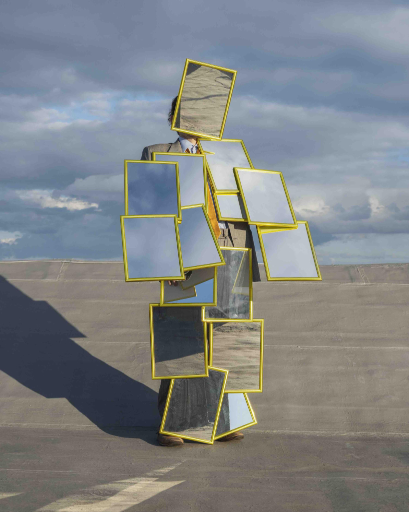 Photo of a figure in a suit standing on grey pavement against a cloudy sky. Their body is almost completely covered in medium-size gold-framed mirrors facing out, stacked from head to toe reflecting the ground and sky.