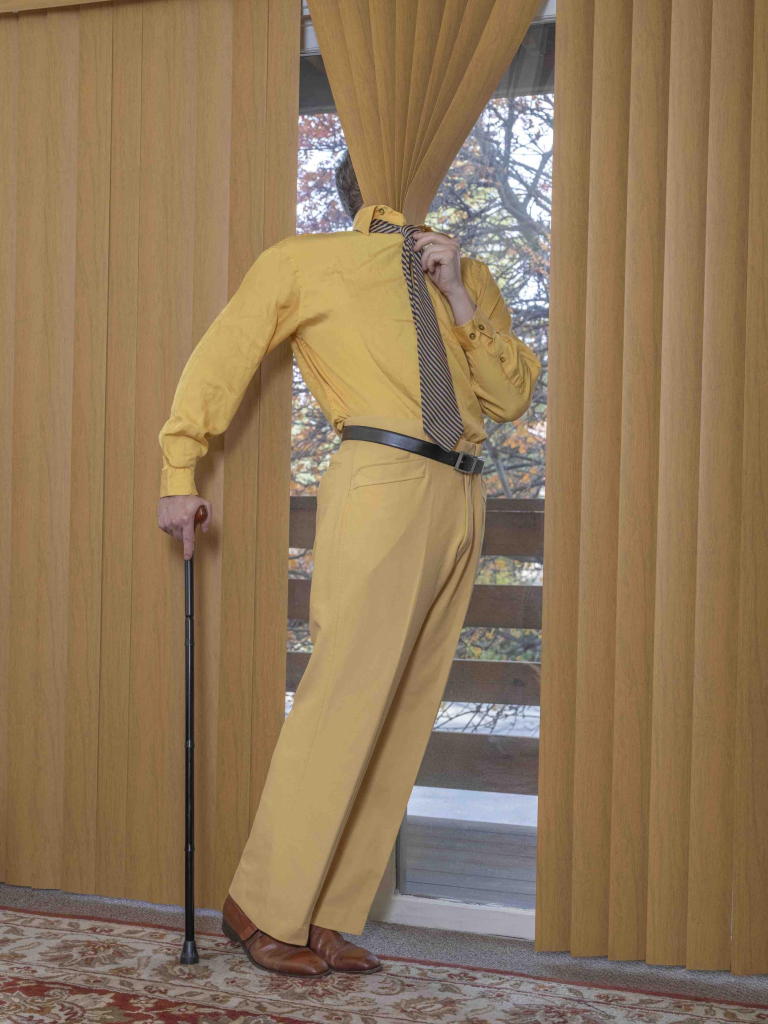 Photo of a white figure in a yellow suit standing near a tall window with long mustard-colored vertical blinds. His right hand holds a cane and his left holding his tie, and his head is obscured by a cluster of blinds over his face and buried in his collar.