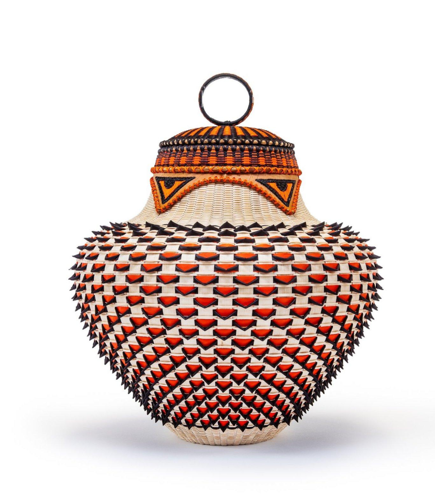 Squat rounded basket with a 3D alternating pattern of red and cream. The lid features several horizontal patterned lines in orange, red, and teal, with two triangle shapes coming down resembling stylized hawk or eagle eyes.