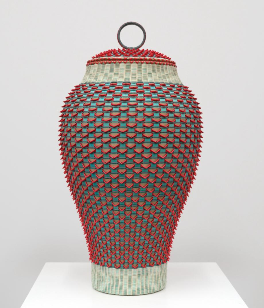 Tall, oblong basket with an alternating 3D grid pattern of red and teal, with pale cream borders at top and bottom and a metal ring over the lid.