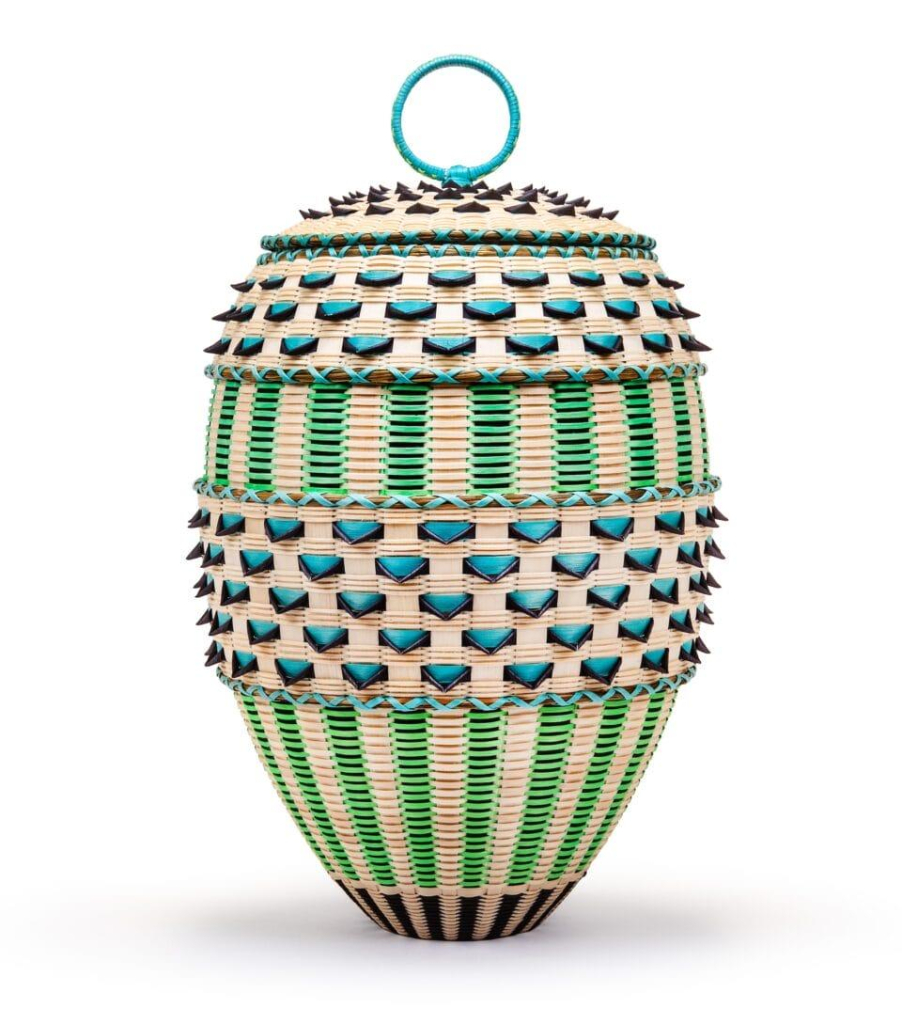 Tall, oblong basket with patterns of green and cream vertical stripes and alternating blue and cream shapes with a 3D effect, and a metal ring over the lid.