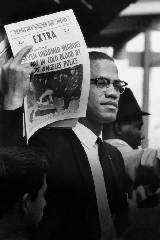 Black and white photo of Malcolm X holding up a newspaper with a headline reading "EXTRA: Seven Unarmed Negroes Shot in Cold Blood by Los Angeles Police".