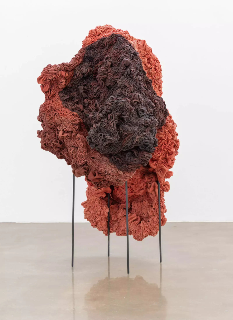 Free standing sculpture of combined red-orange and brown-grey fabrics folded and stitched into a large abstract organic form, hanging over a metal frame with 4 thin legs.