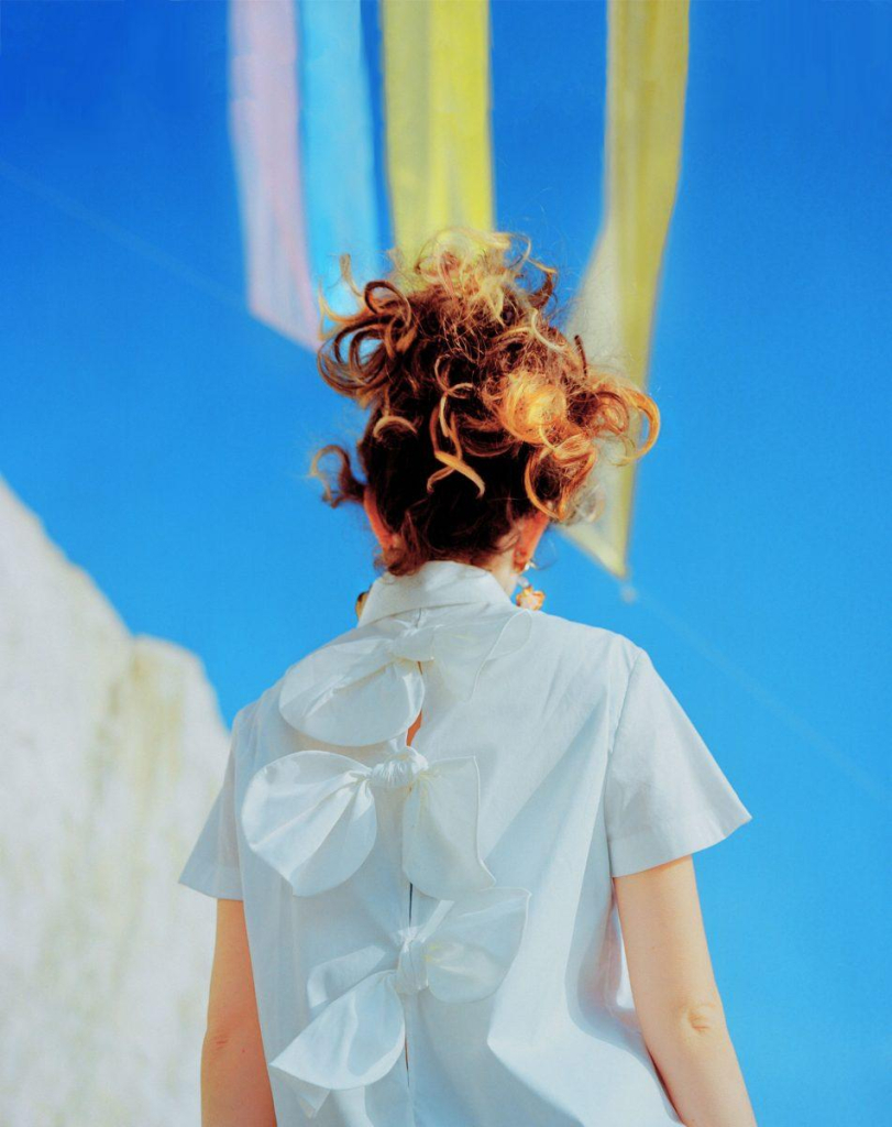 Photo of a white figure seen from behind, with curly red-blonde hair piled on top of their head and wearing a white shirt with large floral-like bows tied up the back. There is a blue sky and white cliff in front of them with colored streamers/banners floating in the sky.