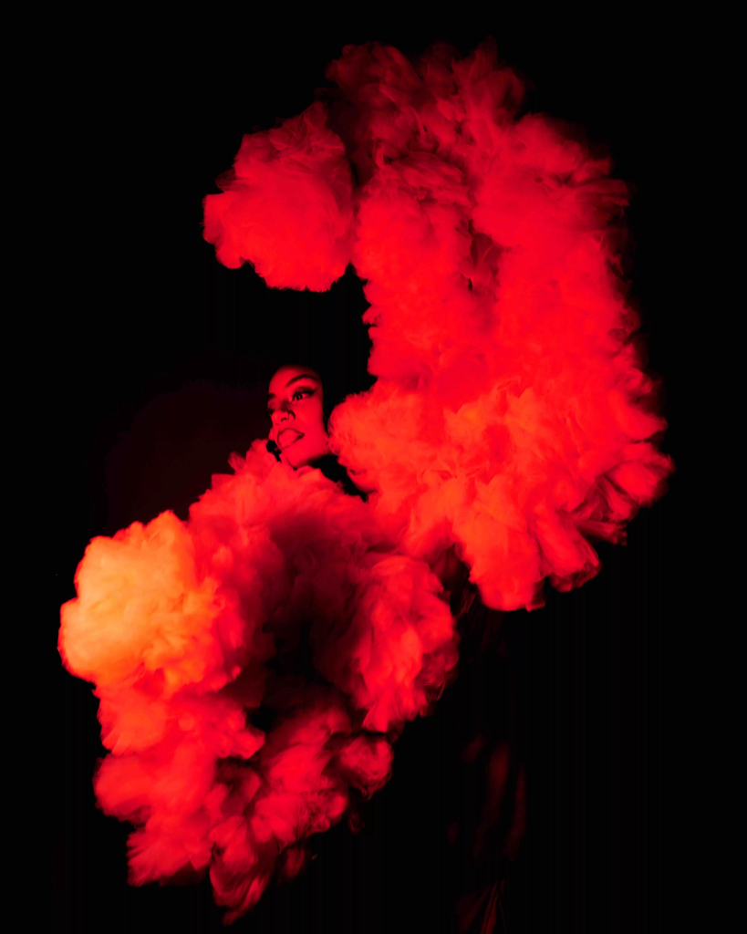 Photo of a Black figure standing in a dark space, surrounded by huge orange-red tulle sleeves framing their upper body and curving up over their head.