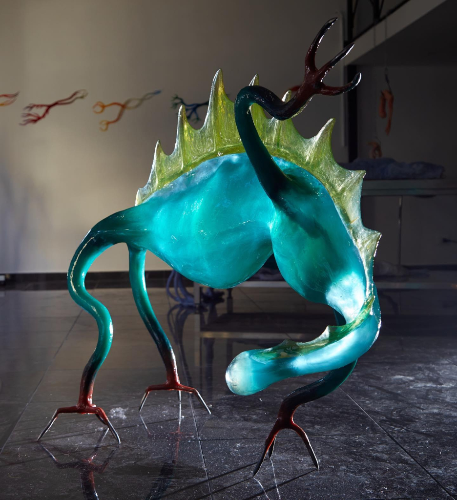 Glass sculpture of an alien creature with a blue body and green back fins, standing on slender red claws.