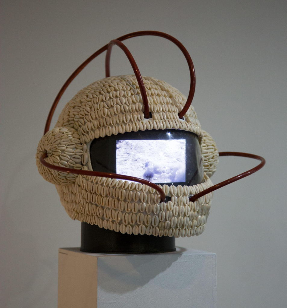 Helmet sculpture covered in cowry shells, with small round protrusions like earmuffs on either side. Inside its visor is a small video screen, and several red tubes emerge to loop around the head.