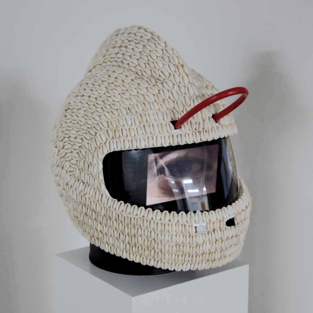 Helmet sculpture covered in cowry shells, with a slightly pointed protrusion along the center top. Inside its visor is a small video screen, and a red tube emerging in a small loop above the visor opening.