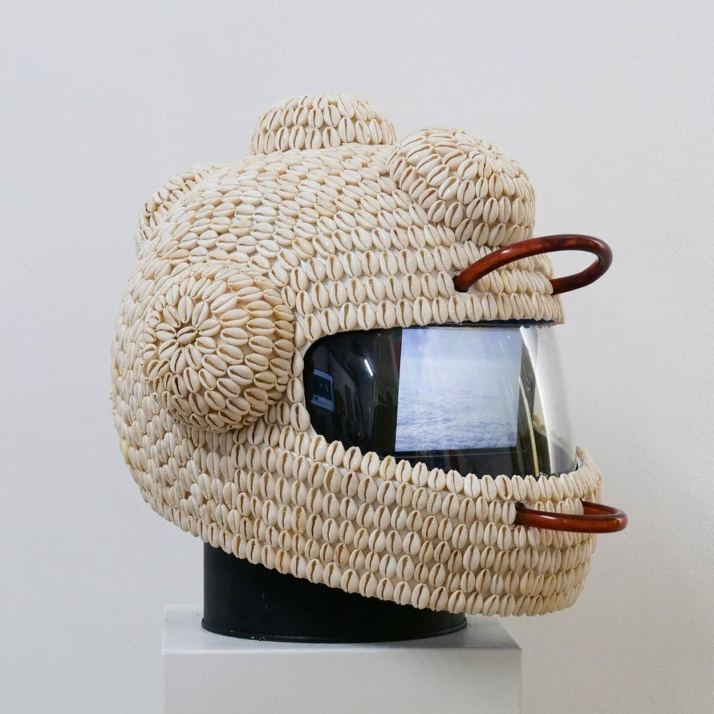 Helmet sculpture covered in cowry shells, with small round protrusions on the sides and top. Inside its visor is a small video screen, and two red tubes emerge in small loops above and below the visor.