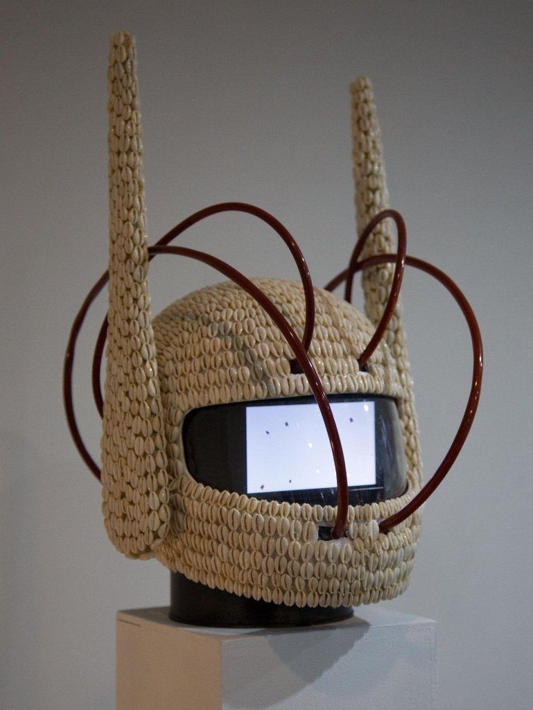 Helmet sculpture covered in cowry shells, with tall wing or horn-like flaps on either side sticking up vertically. Inside its visor is a small video screen, and several red tubes emerge to loop around the head.