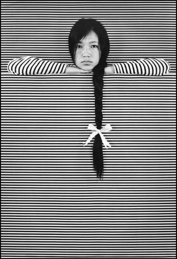 Black and white photo of a Chinese girl wearing a striped shirt and leaning over a striped wall, her long black braid cascading down over the pattern.