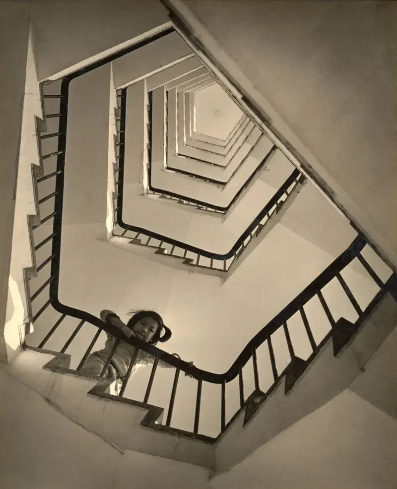 Black and white photo of a Chinese girl leaning over a stair railing, shot from below so that the geometric form of the tall staircase spirals upward in a series of hexagons.