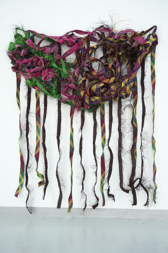 Hanging sculpture of a mass of knotted and overlapping textile strips in green, magenta, and yellow, with various strips falling down to the floor with ragged edges.