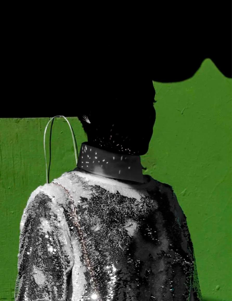 Photo of a Black figure in grayscale wearing a white and grey textured top with their head obscured in shadow, against a green wall.