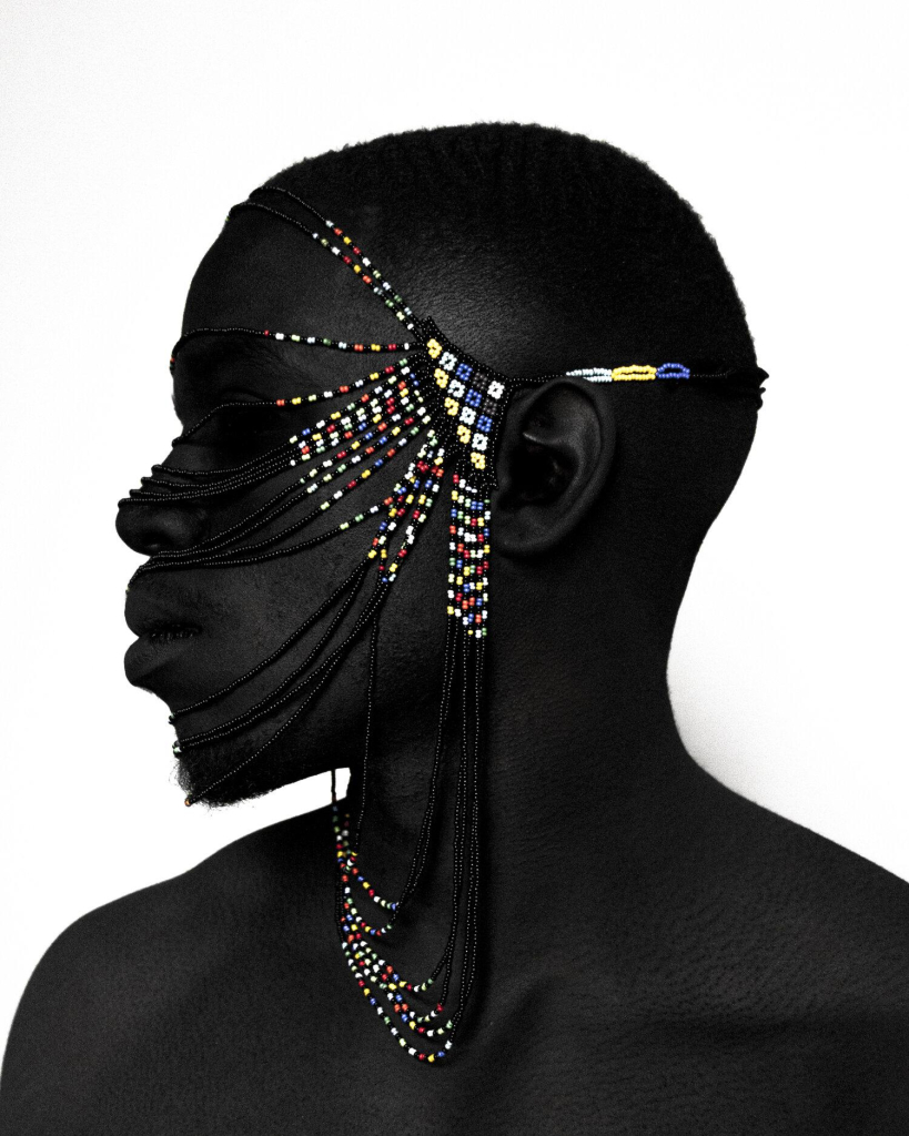 Photo of a Black man in profile wearing a colorful beaded headpiece falling across his face and neck, against a white wall.