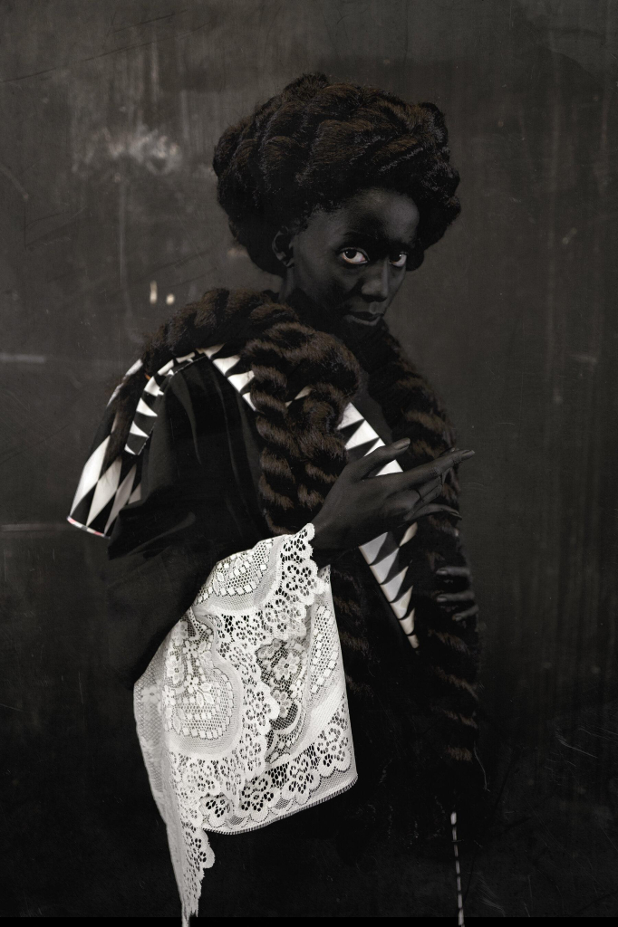 Photo of a Black figure in grayscale wearing a layered outfit of lace and velvet, with hair piled and wrapped atop their head and a thick coiled braid falling down and wrapping around their shoulder, posing against a grey wall.