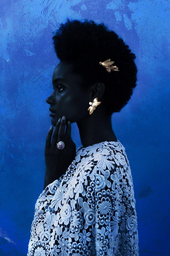 Photo of a Black woman in profile wearing a white floral top and gold butterfly clips in her natural hair, with a hand up to her chin, against a blue wall.