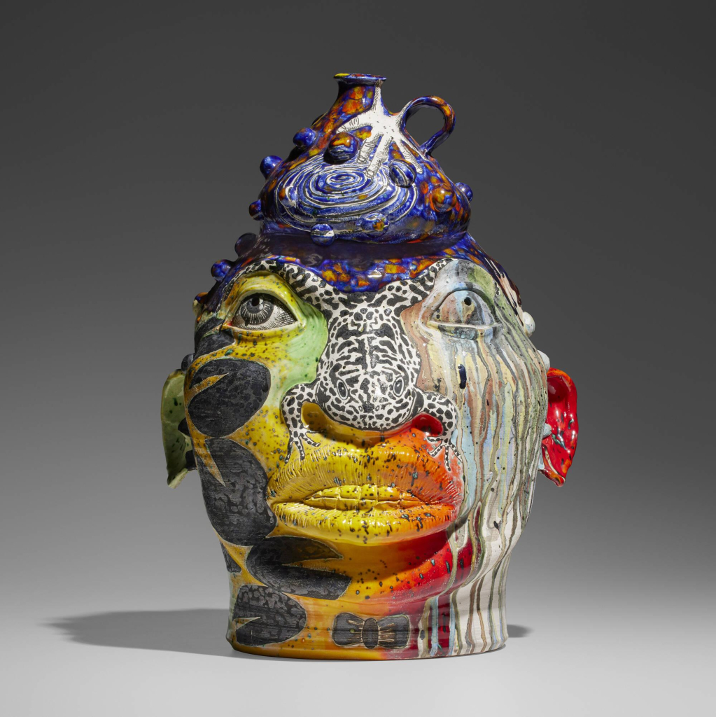 Ceramic sculpture of a human head, with wide eyes and a frog over the nose, covered in various colors and patterns, balancing a vessel on top with a small spout and handle.