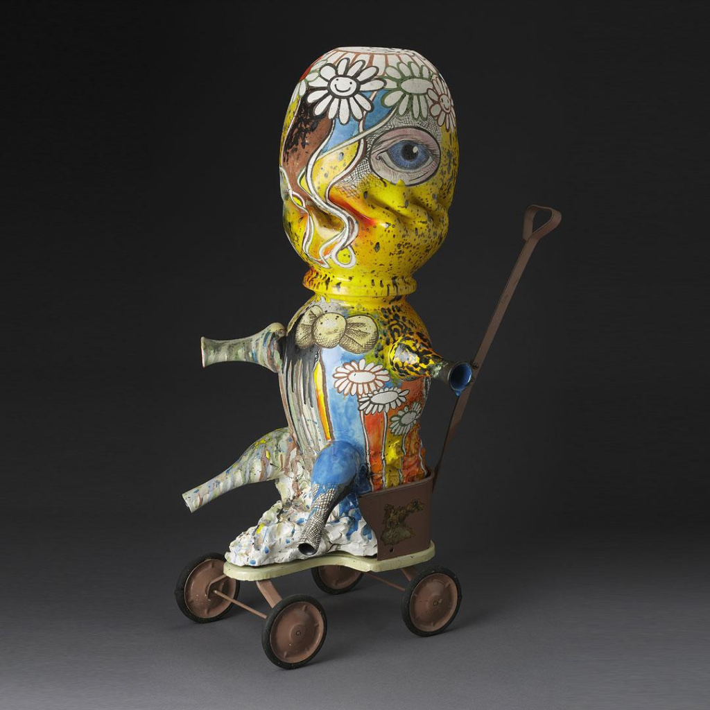 Ceramic sculpture of an abstract figure made up of rounded vessels for the head, body, and limps, sitting on a wheeled platform with a long handle. It is decorated with floral designs and colorful glazes, and a large blue eye.