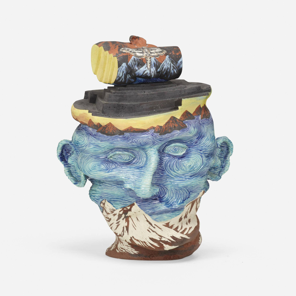 Ceramic sculpture of a lumpy human head decorated with swirling blue and green lines. A painting of a mountain range covers their chin and jaw, and on their head is a another mountain range and a dark grey layered structure.