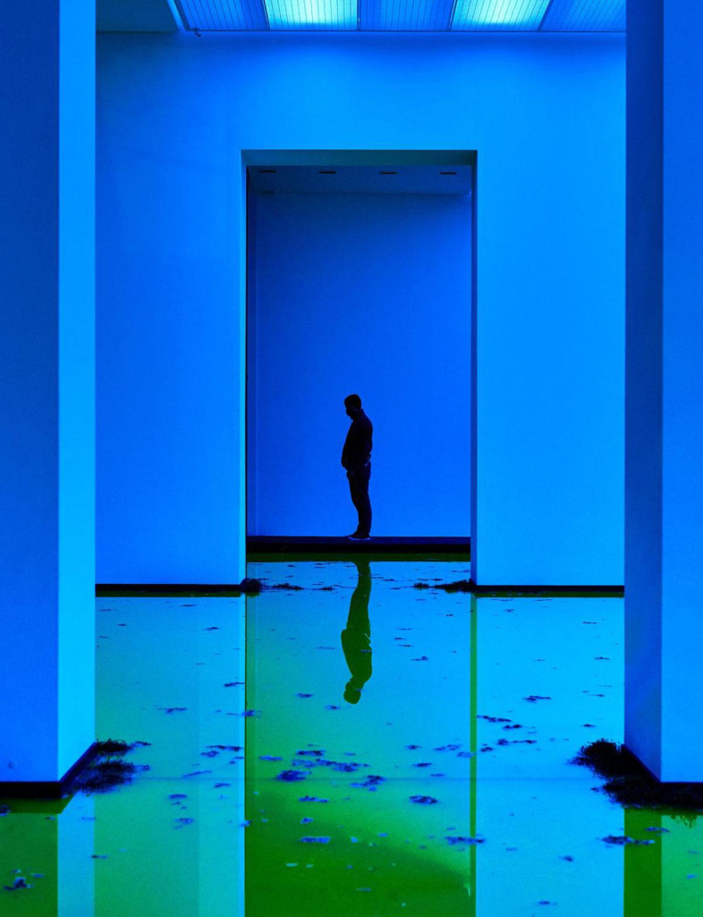 A figure standing at the end of a gallery hallway, with walls lit by blue light and the ground covered by murky green water.