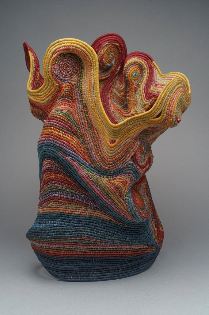 Abstract sculpture of a textured, askew vessel with rounded edges on top, made up of blue, red, and yellow coiled threads.