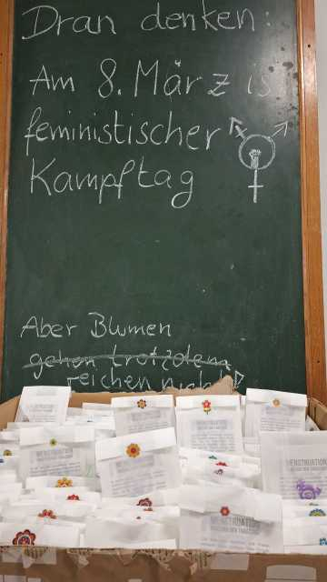 Der Karton mit den befüllten Butterbrottüten vor einer Tafel. auf der Tafel steht: Dran denken: Am 8. März ist feministischer Kampftag. Aber Blumen (durchgestrichen) gehen trotzdem - reichen nicht.