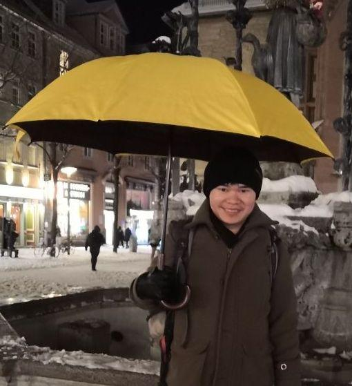 Image shows researcher (Linh Dang) with yellow umbrella standing outside in Göttingen's market place. There is snow around and he is just in front of the Gaenseliesel fountain where we meet.