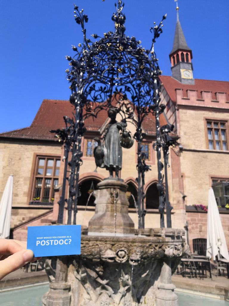 Image of Gaenseliesel Fountain in the Market Place of Göttingen. It has a decorative wrought iron all around her. There is someone holding a blue card that says "what would you change as a postdoc?" on it