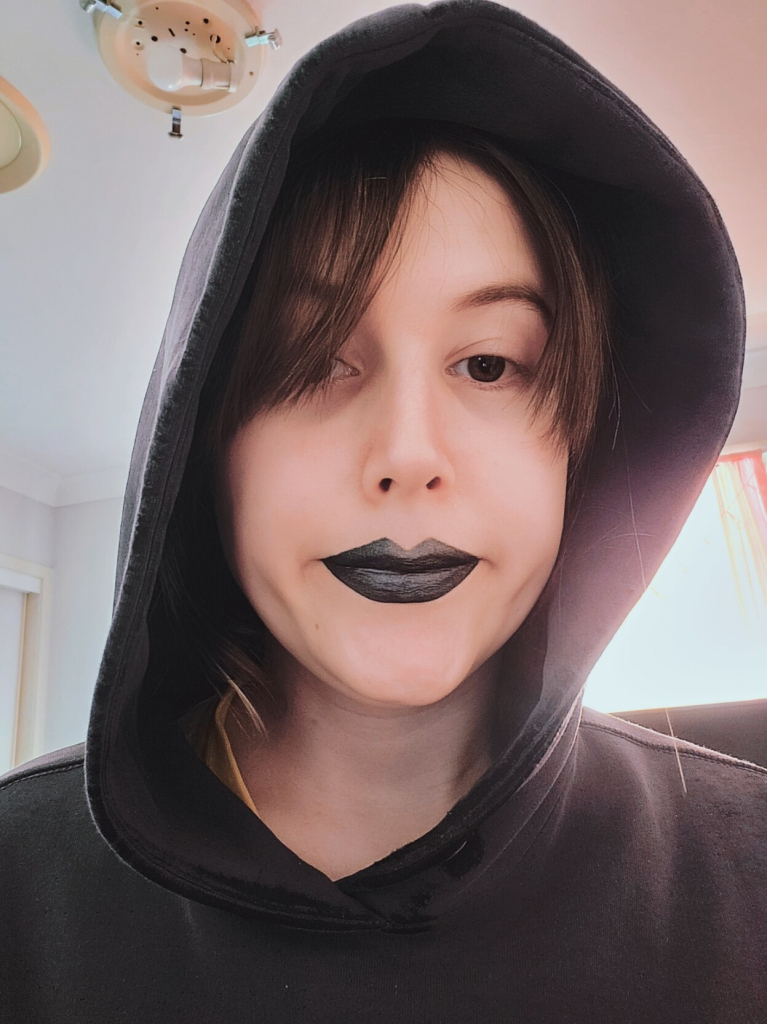 Selfie of Revy
She's wearing a black hoodie
Black lipstick with a silver tone
And is slightly smiling
While her short brown hair covers on of her eyes