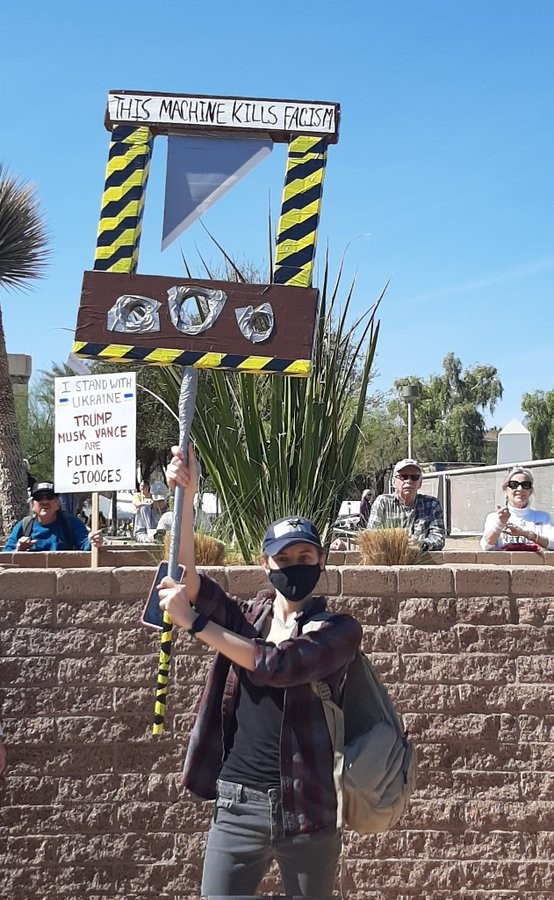 Picture of a protester holding a placard in the style of a guillotine, with the words THIS MACHINE KILLS FASCISM written across the top.