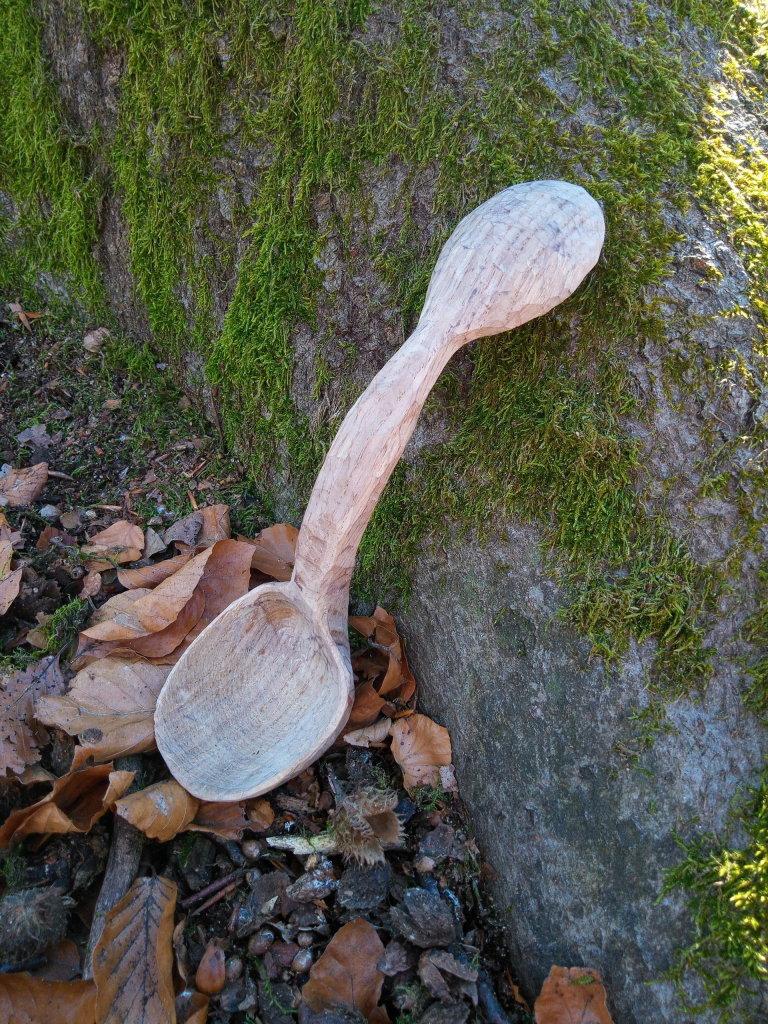 Ein geschnitzter Doppellöffel ist an einen Baum mit grünem Moosbelag angelehnt, der tiefe Schlürflöffel ist unten.