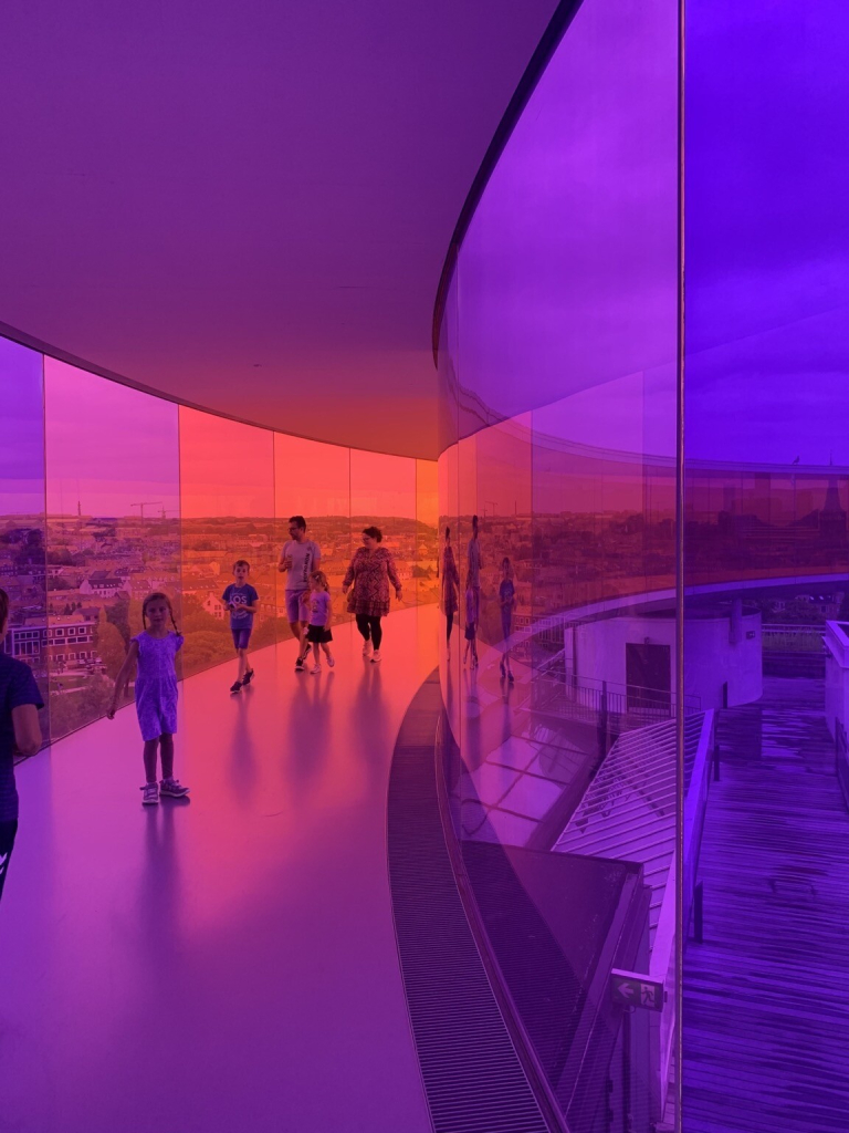 A family walking around a circular walkway on the roof of an art gallery. The windows are tinted with different colours all the way round, here merging from blue to purple to pink to orange.