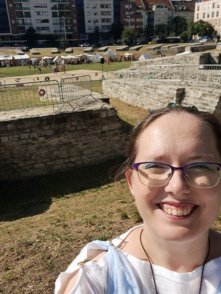 Selfie with the ruins of the Amphitheater in the background. I (30s white woman, red hair, glasses) am wearing white and blue Roman garb. Behind me in the arena there are tents and yurts for the reenactors, and a large square of sand for the upcoming gladiator fights.