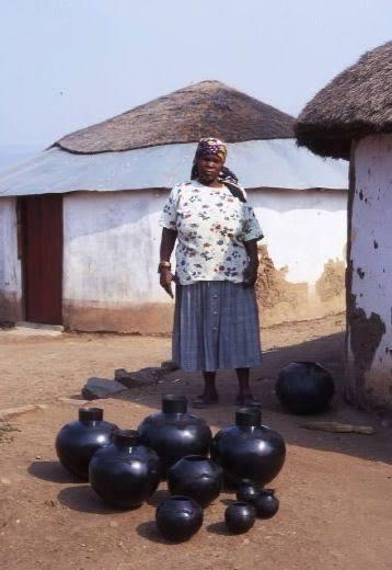 A photo of Zulu potter Nesta Nala (1940-2005) standing behind a batch of her shiny black pottery. 