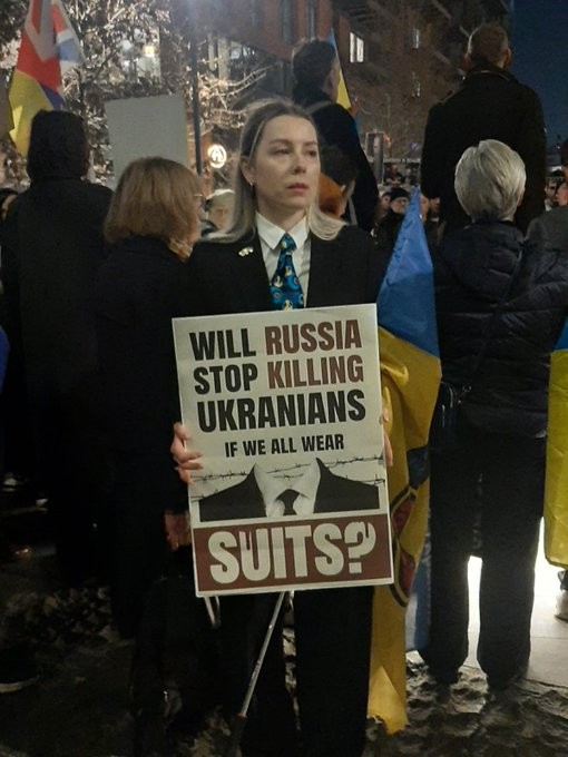 Woman in a suit holding a placard reading
WILL RUSSIA STOP KILLING UKRANIANS IF WE ALL WEAR SUITS?