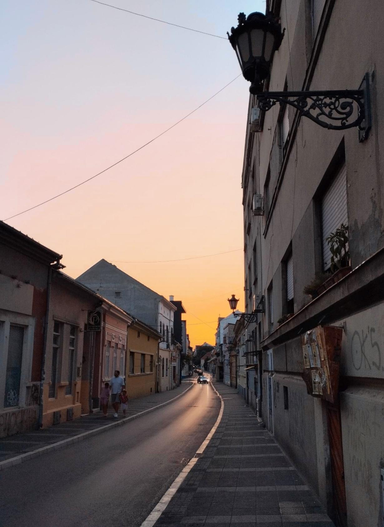 A narrow street with small houses to the right and left in the warm orange-coloured, darkening light of the setting sun on the horizon.

Eine schmale Straße mit kleinen Häuschen rechts und links im warmen orangefarbenen, dunkler werdenden Licht der untergegangen Sonne am Horizont.