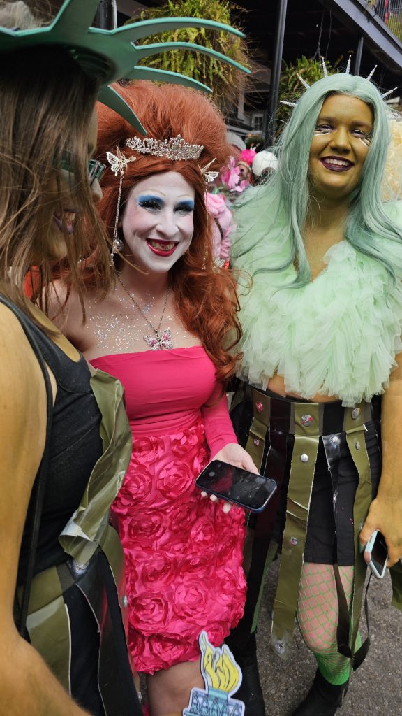 Three people in costume, two are facing the viewer. The first one has large puffy red hair, with a tiara and butterfly decorations, and a hot pink dress with ornate 3 dimensional rose design. The second one has light green hair, with decorative spikes in semicircle, and same color feather shoulder/chest piece. Under that is a skirt with moss green and black vertical straps with metal details. 