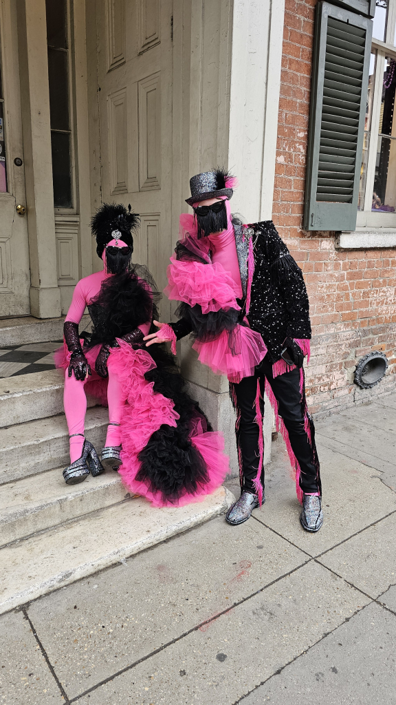 Two people in costume and masks. Both are in black and hot pink. Both are wearing black fringe face covers. One on right has a glittery black suit with pink fringe accents and a glittery black top hat with a pink feather. Very glittery black shoes. On the left side is a large accent of pink bunched sheer fabric longer than the person's torso. 

The other on left is sitting on concrete steps with hot pink body suit, pink and black tutu, black gloves, bunched black sheer fabric front that extends to long train in pink and black, curled to the front on the stairs. Very glittery black pumps.

The background is portion of historical building in New Orleans French Quarter. 