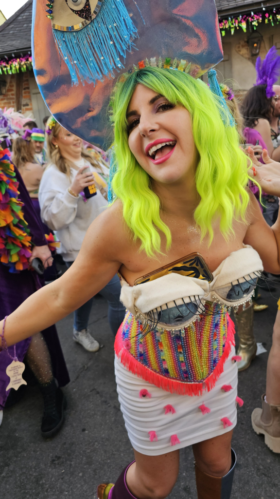 Woman in costume in front of a crowd of people on a street. She is posing for the camera, and has a large disc headdress, blue and pink, with a light-blue fringed eye image, bright green hair. She is smiling with her mouth open left and right arms extended out and back. Her costume includes two detailed blue eyes covering her breasts. A phone is tucked behind one of these eyes. Below these eyes are vertical stripes in bright colors with included geometric shapes in contrasting colors, ending in hot pink fringe at hip level. The skirt is tight white with horizontal accents. 