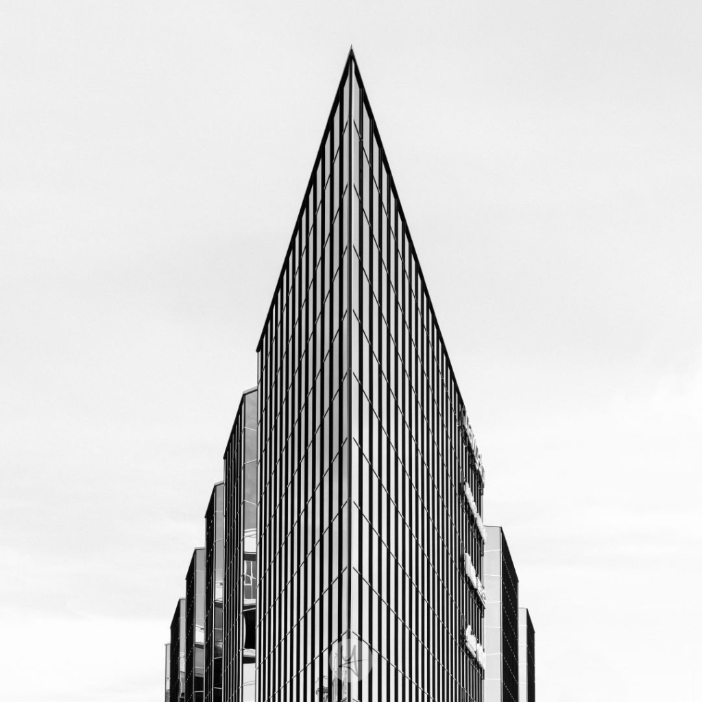 Black and white photo of a triangular shaped office building. The perspective is on the very pointed edge of the building, looking up, so that it nearly looks like a pyramid. The face of the building is made out of glass with lots of windows und only divided by its metal structure.